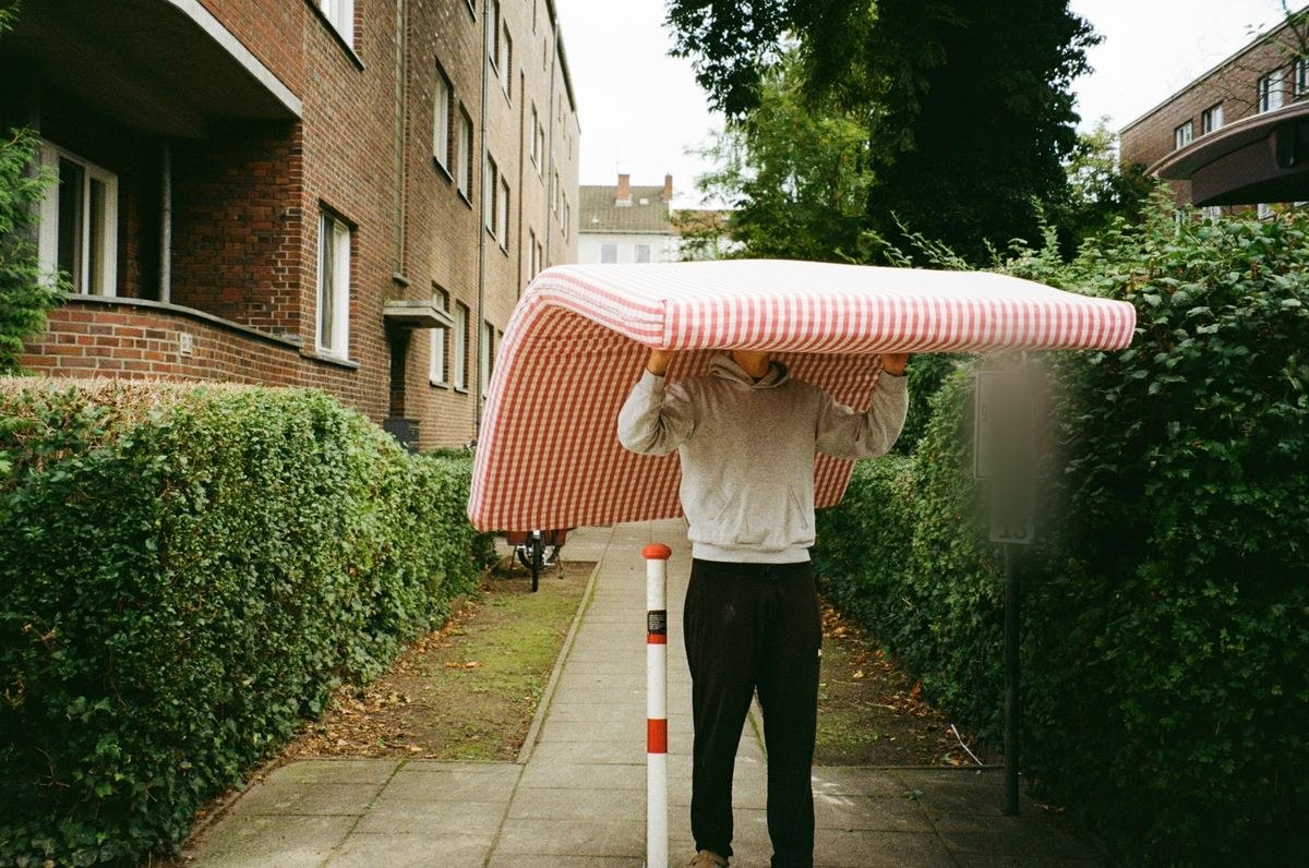 Iemand draagt een matras over het hoofd op een tuinpad tussen twee hagen