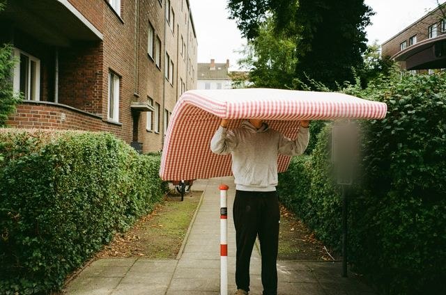 Iemand draagt een matras over het hoofd op een tuinpad tussen twee hagen